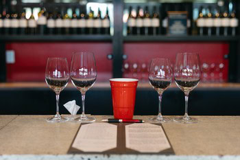 Four empty wine glasses and a red cup on a bar counter with a wine shelf in the background.