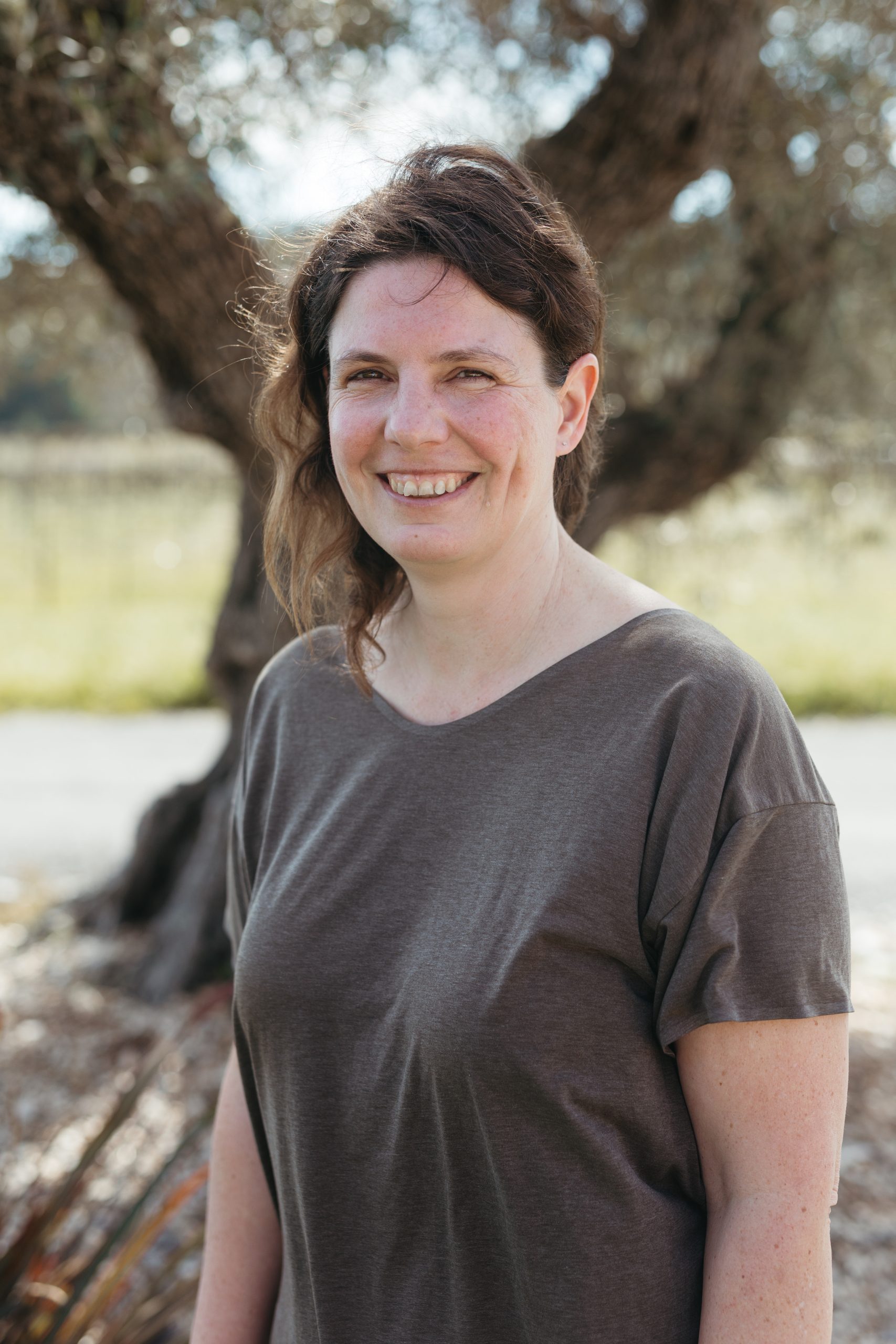 Smiling woman outdoors near a large tree, casual gray t-shirt, natural background.