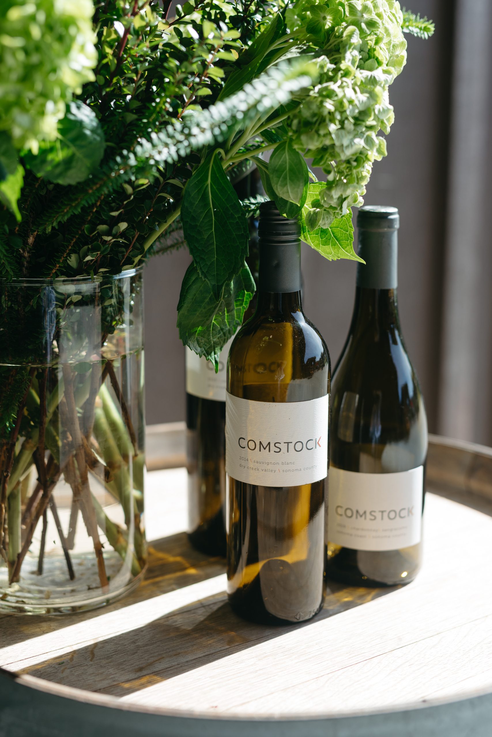 Two bottles of Comstock wine on a wooden tray with a floral arrangement in the background.