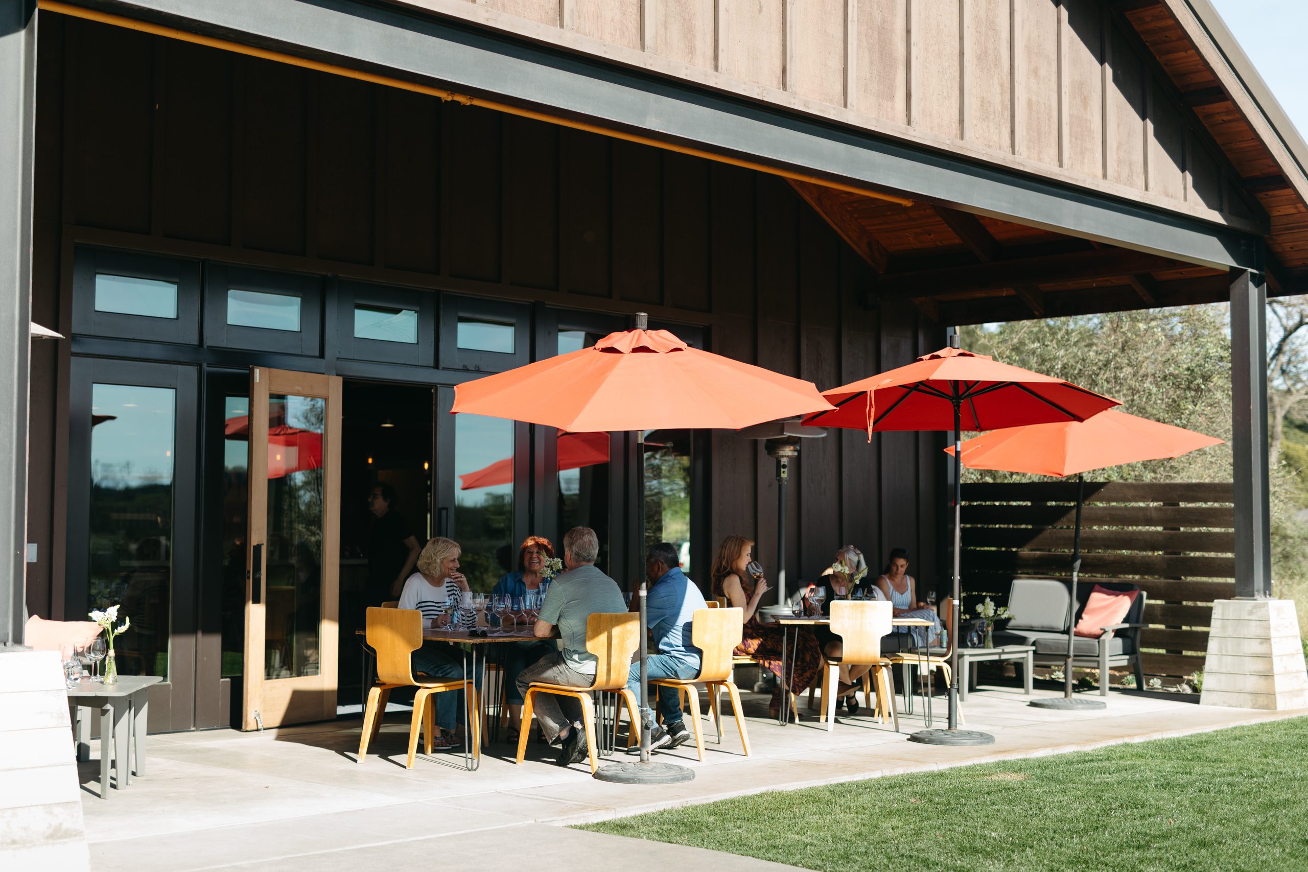 Outdoor dining area at Comstock Wines with guests under red umbrellas on a sunny day.