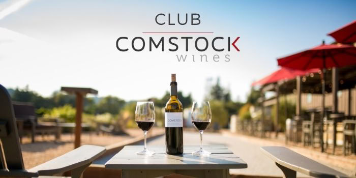 Two wine glasses and a wine bottle on a table outdoors at Comstock Wines, with a vineyard background and red umbrellas.